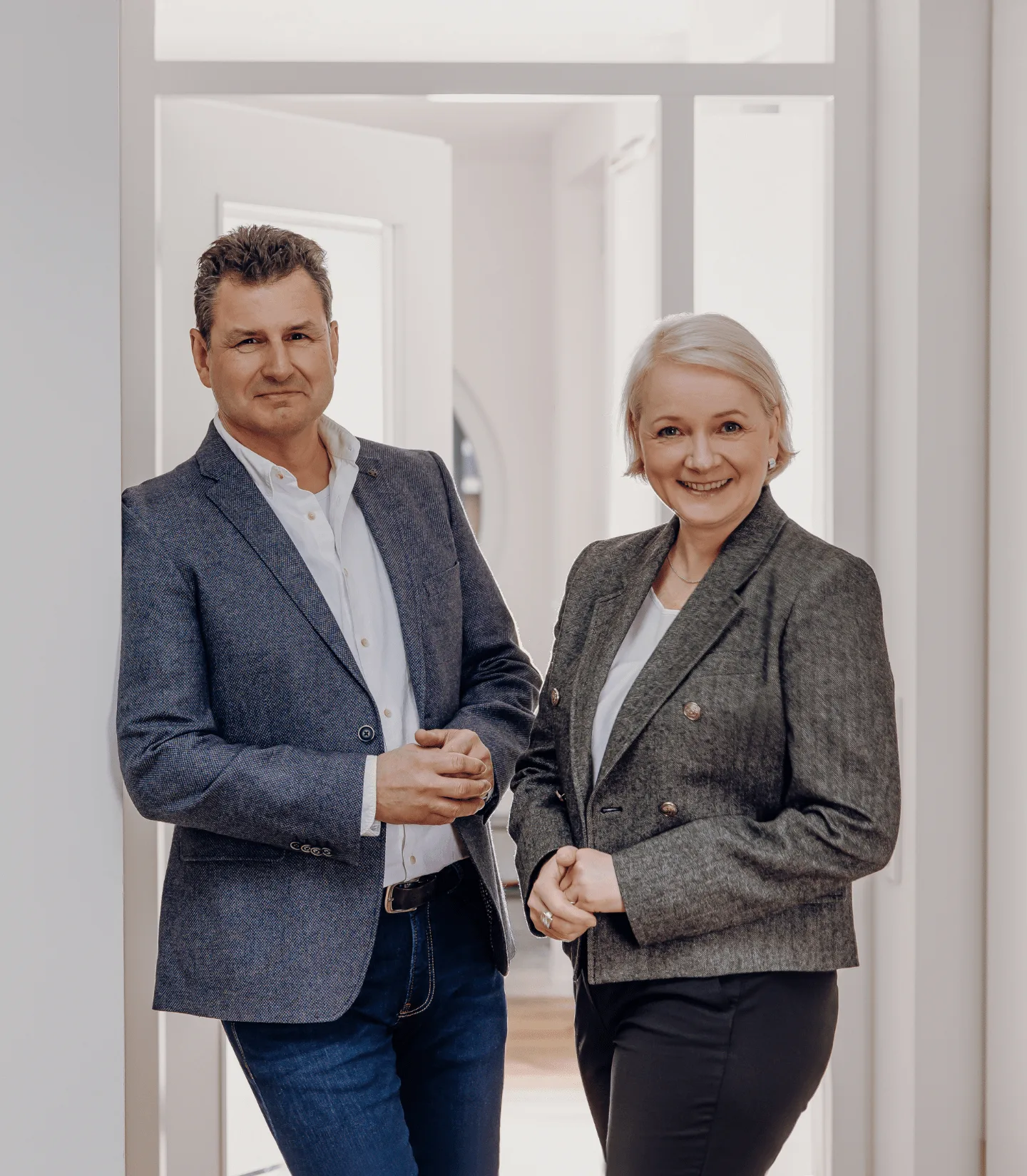 Zwei Personen in hellen Hemden posieren lächelnd an einem dunklen Tisch in einem modernen Raum mit grüner Wanddekoration.